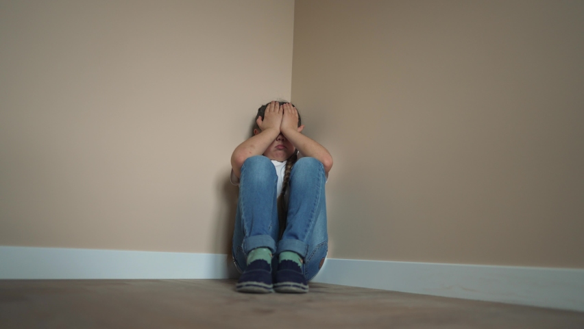 School-age child is sad in corner of room. Sad, crying child offended. girl is sitting in corner on floor of room indoors. Punishment of children at home in family. Brutal upbringing at home