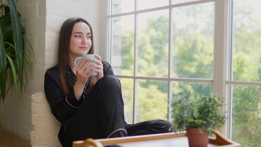 Side view of woman in pajamas drinking coffee in morning and looking out window, slow motion. Greenery on background. Girl relaxing and enjoying
 Royalty-Free Stock Footage #1092918869