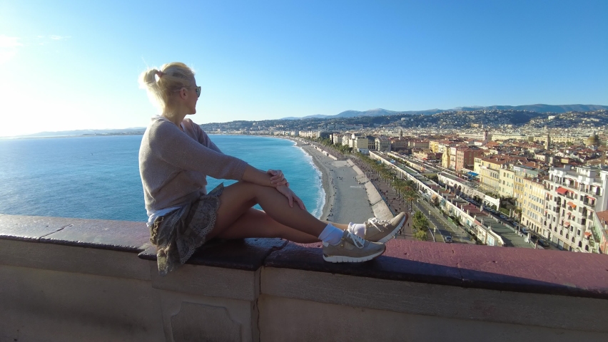 Girl sitting at overlook of French city skyline of Nice on des Anglais promenade. Skyline of Nice with Ponchettes beach from panoramic terrace of Colline du Chateau park in Provence of France.