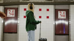 Female tourist traveling with a suitcase on the edge of a platform. Blonde-haired passenger in black headphones dancing at the subway station. High quality 4k footage