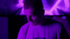 DJ in club working indoors in neon lights close up. Caucasian male deejay person in nightclub using headphones, laptop and control console to mix music tracks. Nightlife concept. Slow motion 4K video