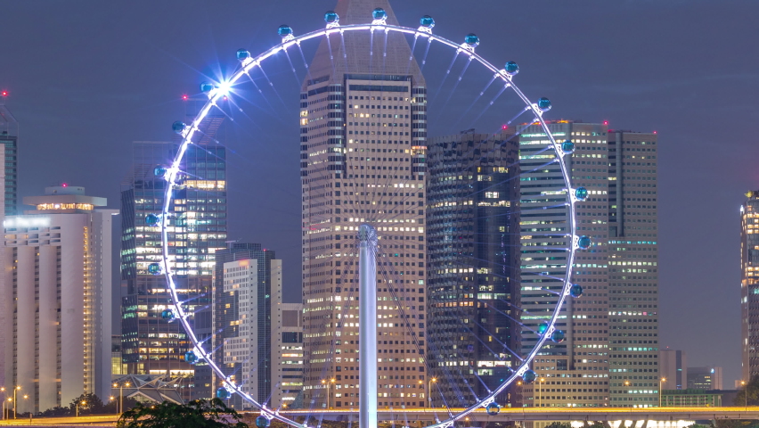 Skyline of Singapore with famous Singapore Ferries Wheel night to day transition timelapse. Illuminated skyscrapers on a background before sunrise