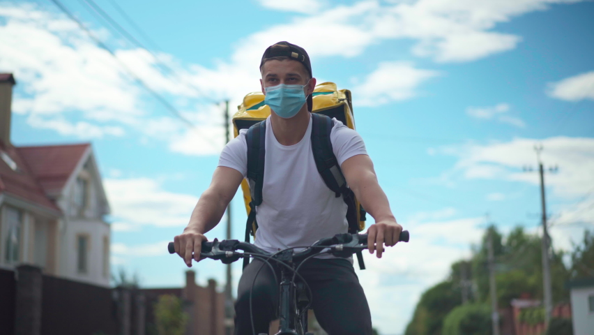 Front view portrait of young man in coronavirus face mask riding bicycle delivering food and drink in urban city. Confident Caucasian delivery boy driving bike with backpack at background of sky | Shutterstock HD Video #1092943715