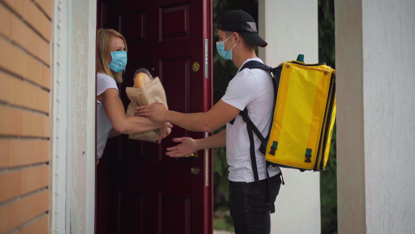 Courier in Covid-19 face mask knocking in door as positive woman opening taking food delivery. Side view Caucasian young man and woman passing parcel outdoors at house entrance | Shutterstock HD Video #1092943729