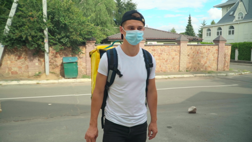 Positive delivery boy in coronavirus face mask passing parcel to client at gate in town. Portrait of Caucasian young man delivering food and drink on Covid-19 pandemic outbreak | Shutterstock HD Video #1092943739