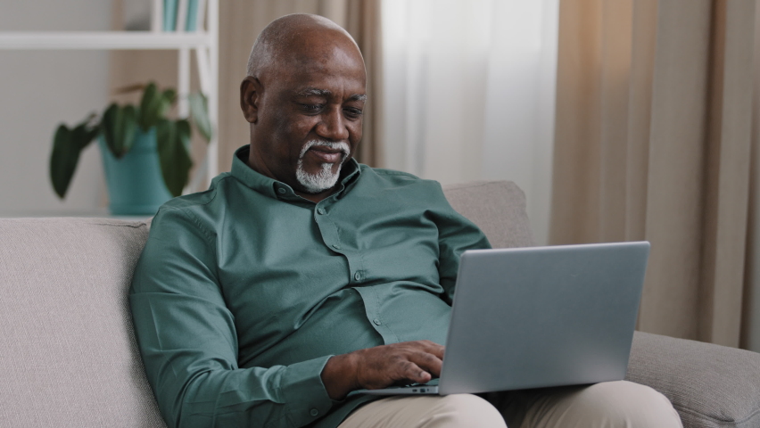 Older bald African American man 60s senior mature businessman use easy wireless technology for comfort work online shopping ecommerce at home couch typing massage chatting banking in computer app Royalty-Free Stock Footage #1092944891