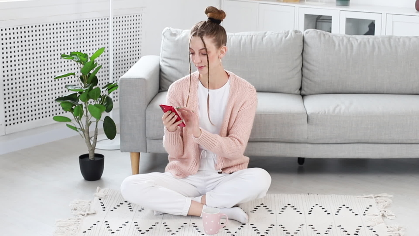 woman smiling in the living room on the floor with a smartphone and drinking from a cup