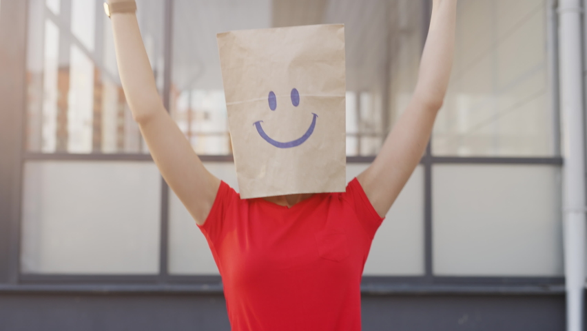 A crazy anonymous woman is gesturing with a cardboard box on her head with smiley face. Funny person is doing winner gesture and celebrating. Concept of victory or happiness and freedom.