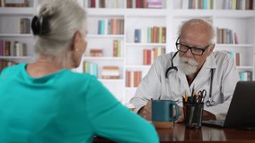Mature senior male doctor talking to female patient in modern clinic with books background.