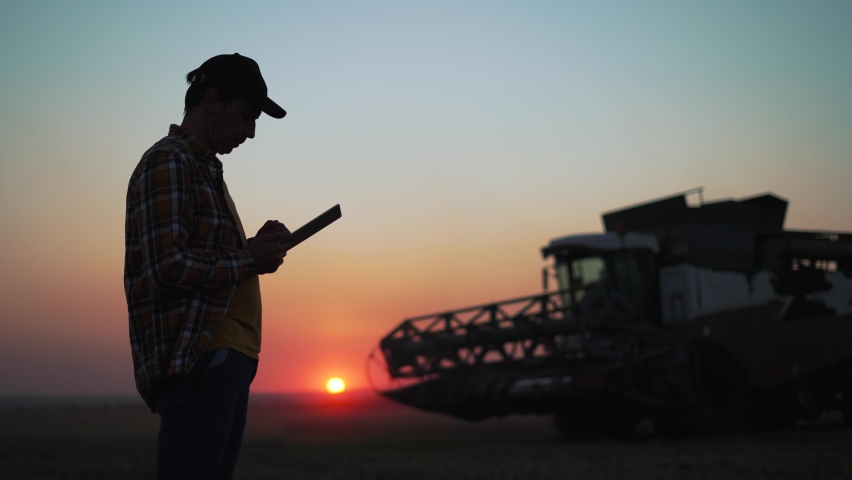 Agriculture. Harvester harvests wheat field at sunset. Silhouette of farmer with tablet in wheat field.Harvesting at sunset.Agricultural business.Farmer controls harvesting of wheat grain by combine Royalty-Free Stock Footage #1092956181