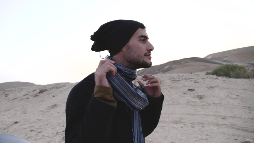Jewish man traveler standing by the tent swinging scarf around the neck while at sunset in Ramon Crater Trail, Negev desert, Israel in slow motion
