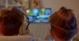 The backs of the heads of children in front of the TV. They are holding video game controllers in their hands. On screen in bokeh action game. Spending free time on holidays.
