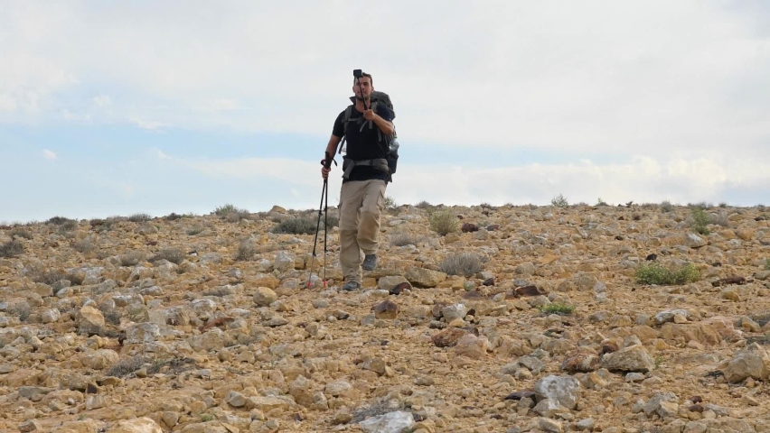 Low angle shot of a hiker talking on a live video while walking on Mount Ramon desert trail on the way to Ramon Crater in the Negev Desert, Israel at daytime.