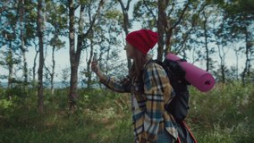 Woman traveller on hike in epic forest. Happy and excited young woman in adventure gear and with a backpack on her back walks through the woods overlooking the seascape and talks on a video call