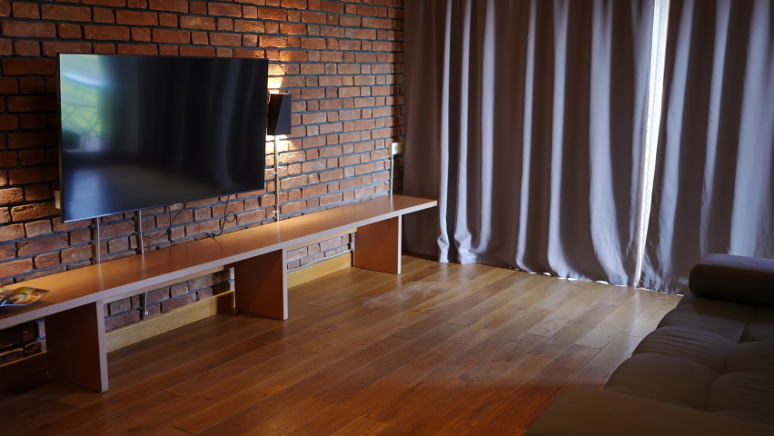 Big modern TV-set hanging on brick wall with comfortable couch in hotel room. Large window closed with curtains. No people indoors at tourist resort. Comfort and service concept