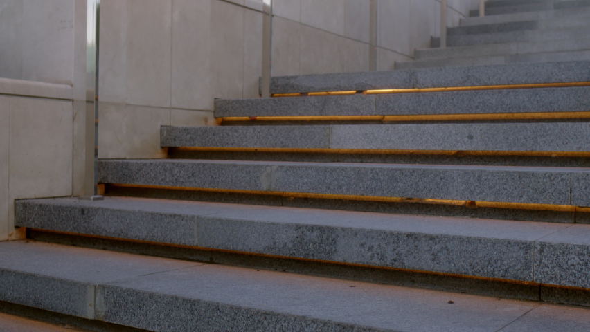 Unrecognizable young businessman climbs up backlit stone stairs in city. man in formal suit dark blue color with tablet in his hands walks forward, close up view of his feet in brown leather boots.