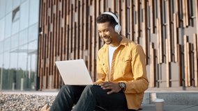Portrait of joyful young man wearing white headphones sitting outdoors and looking at laptop screen watching video. Young male works on computer outside. Remote work, support, call center worker