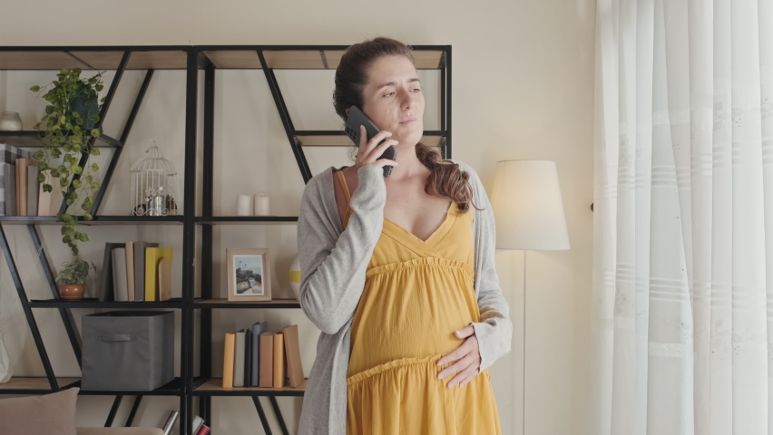 Pregnant woman standing in living room at home, looking through window and speaking on mobile phone