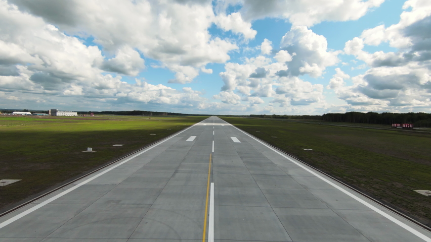 Empty asphalt runway with white markings and light equipment by sides in green field under sky with fluffy clouds