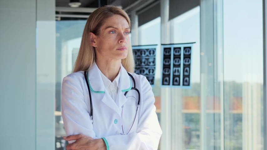 Physician is thinking. Portrait of Young Adult Female professional Doctor standing in modern hospital near panoramic window looking away pensive about problem, wearing lab coat stethoscope indoor.  Royalty-Free Stock Footage #1092984261