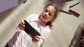 Top View Home: Happy Little Girl Using Smartphone While Lying on the Floor. Joyful Sunny Day and Clever Smiling Child Playing Smart Video Games, Using e-Learning Apps on Mobile Phone. Zoom Out Shot