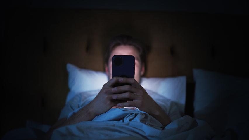 Caucasian Man Uses Smartphone in Bed at Home at Night. Handsome Guy Browsing Social Media, Reading News, Doing Online Shopping, Chatting with Friends Late at Night. Focus on Hand Holding Mobile Phone Royalty-Free Stock Footage #1092986345