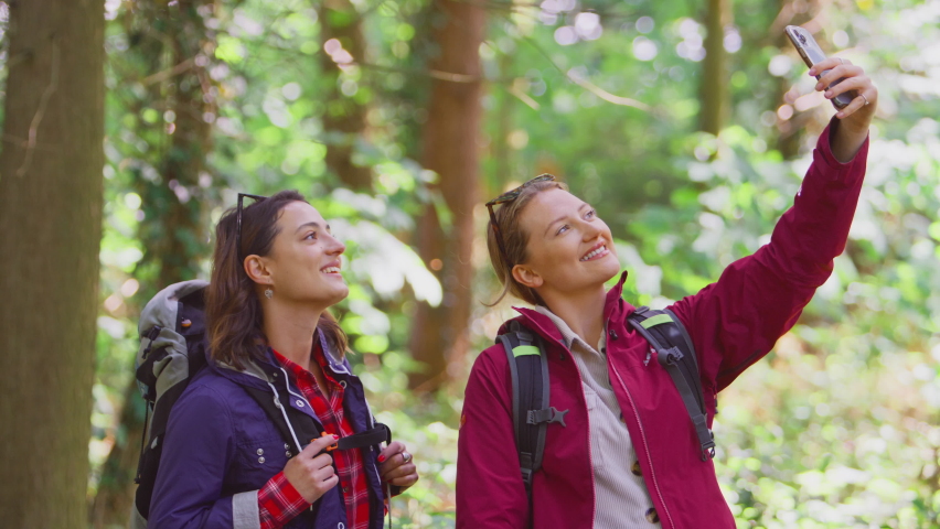 Two female friends posing for selfie on mobile phone on hike through woods together - shot in slow motion