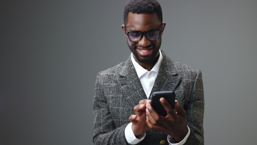 African american businessman man holding a phone in his hands looks at him and is happy to win and win in the office on a gray background | Shutterstock HD Video #1092991067