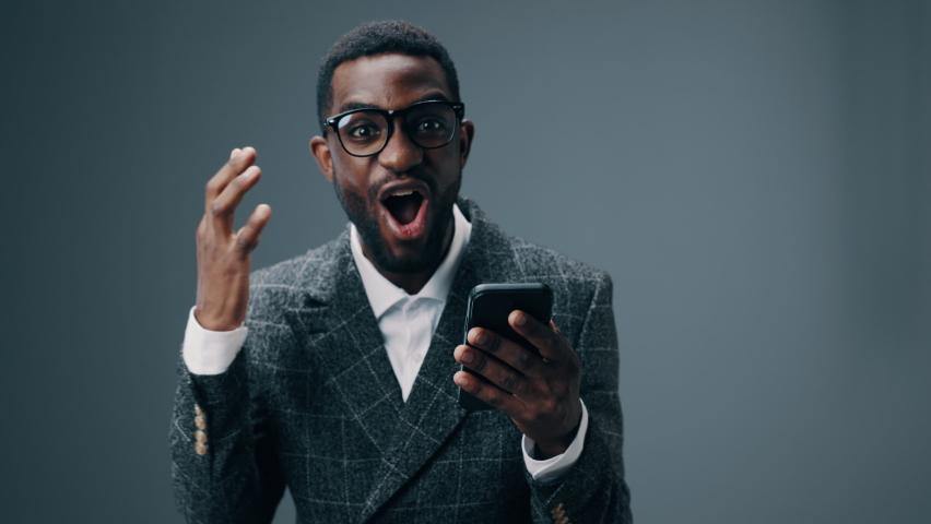 African american businessman man holding a phone in his hands looks at him and is happy to win and win in the office on a gray background | Shutterstock HD Video #1092991073