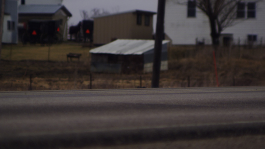 Hooves and wheels of a horse drawn Amish buggy traveling a two-lane blacktop county road pass a bustling Amish farmstead on a gloomy day in Iowa. 