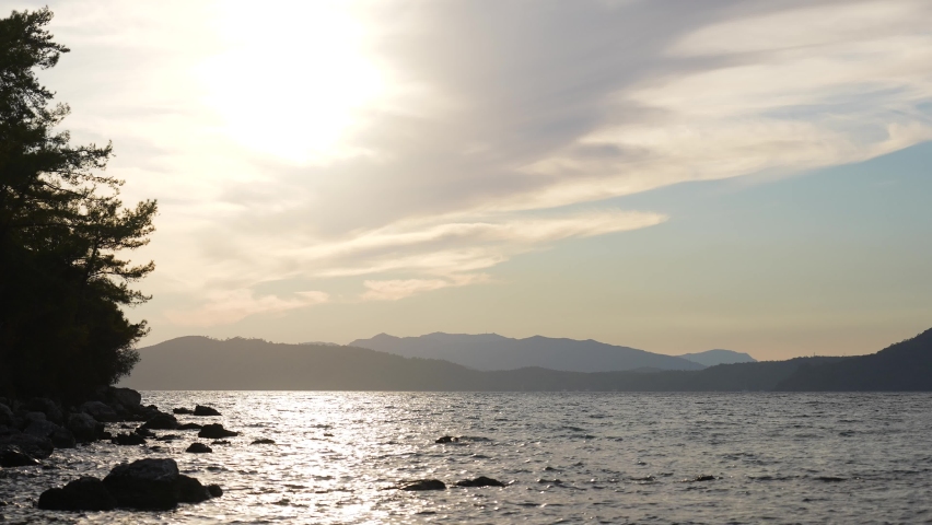 Sunset marine landscape. 4k stock video footage of beautiful peaceful dark evening sea water splashing, silhouettes of green trees, black mountains on horizon line, blue sky with white clouds and sun