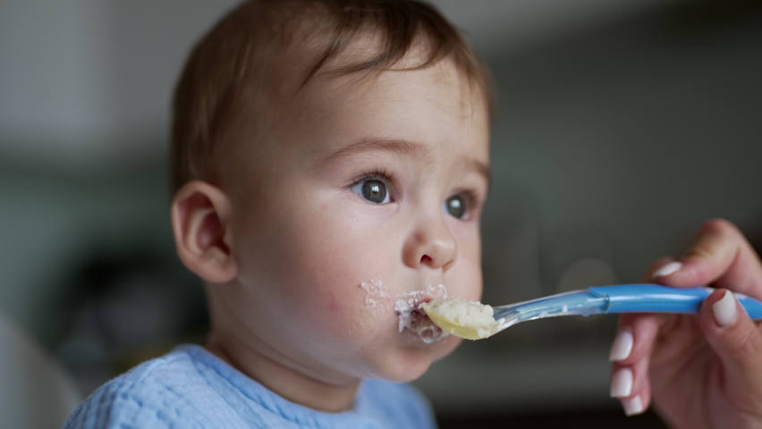 Little kid with adorable plump cheeks eats porridge. Female hand gives a spoon to a baby and cleans child's mouth. Close up. Royalty-Free Stock Footage #1092994917