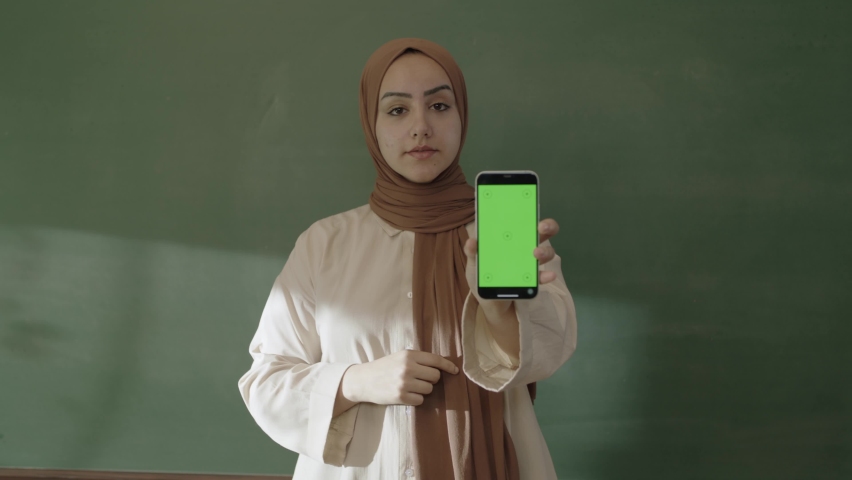 In the classroom,the teacher in hijab looks at social media accounts with her smartphone,browses the internet,reads e-mails,surprised by what she sees and showing her green screen phone to the camera. | Shutterstock HD Video #1092995183