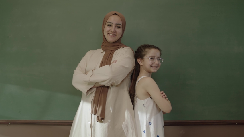 Happy hijab dressed female teacher and little schoolgirl in classroom with arms folded in front of blackboard and looking at camera. Teacher and successful student smiling in front of blackboard.   | Shutterstock HD Video #1092995305