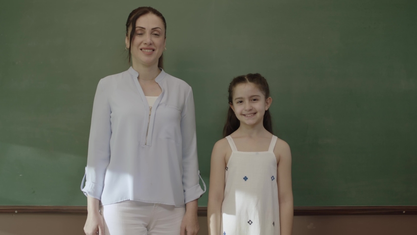 Happy teacher and little schoolgirl standing in front of blackboard in classroom with folded arms and back to back looking at camera. Teacher and successful student smiling in front of blackboard.