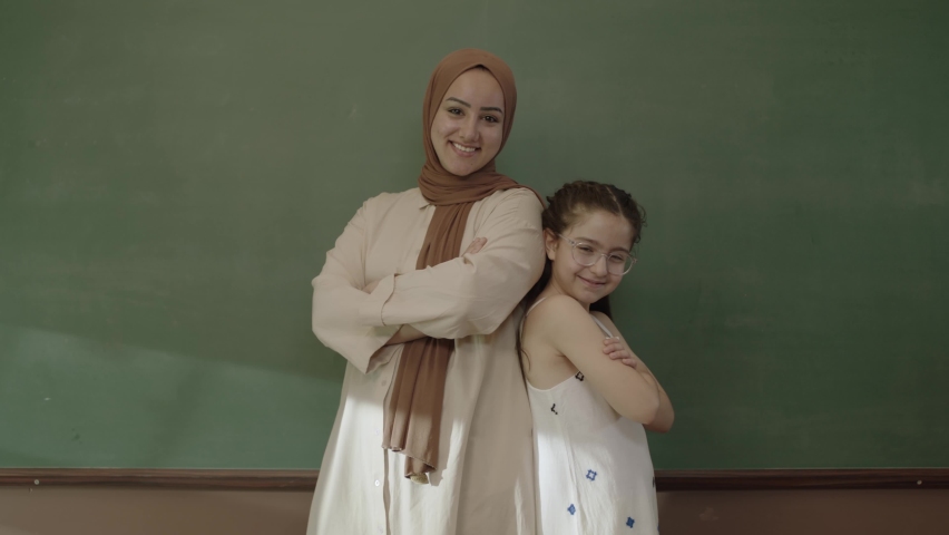 Happy hijab dressed female teacher and little schoolgirl in classroom with arms folded in front of blackboard and looking at camera. Teacher and successful student smiling in front of blackboard.   | Shutterstock HD Video #1092995309
