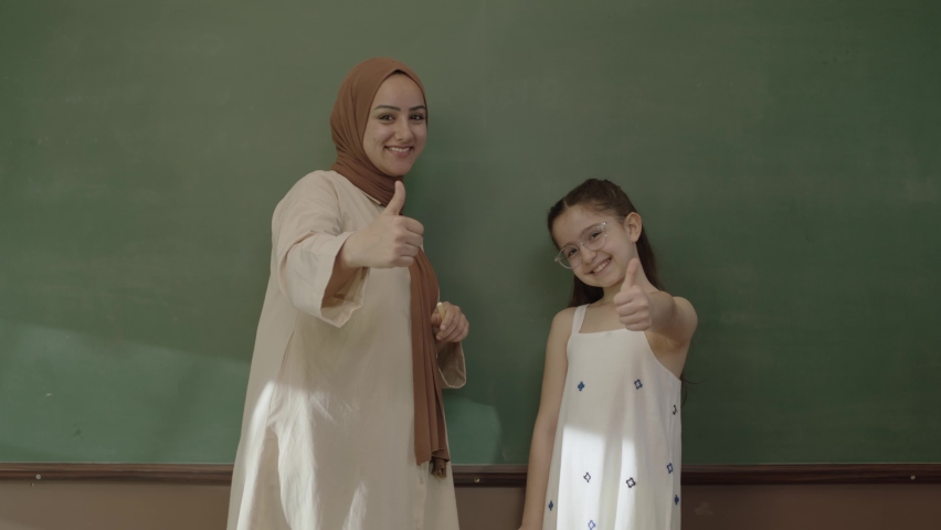 Happy hijab dressed female teacher and little schoolgirl in classroom with arms folded in front of blackboard and looking at camera. Teacher and successful student smiling in front of blackboard.   | Shutterstock HD Video #1092995315