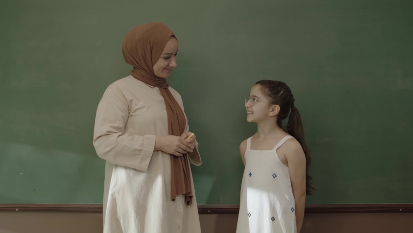 The teacher in hijab teaches the student in front of the blackboard.Little schoolgirl wearing hijab responds to chemistry teacher at the blackboard and writes chemical formulas.Education for children. | Shutterstock HD Video #1092995317
