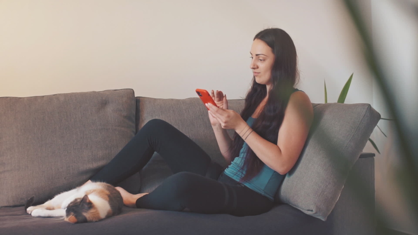 Young woman sit on sofa with cat at home and using smartphone 