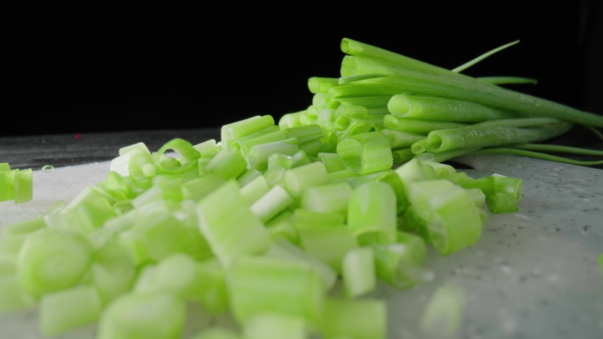 Pieces of green onion stalks chopped on a gray plastic board. Slices of fresh green scallion on a black isolated background. Chopped shallots or chive. Macro shot. Slow motion ready 59.94fps.