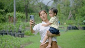 Family recreation and weekend leisure in park. Asian mother makes a selfie with her little son in Green Summer Park, Mom taking photos using mobile phone, They gladly smile at the camera. 