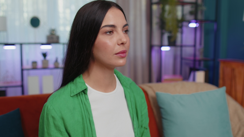 Please, God help. Girl praying sincerely with folded arms, looking upward and making wish, asking with hopeful imploring expression, begging apology. Young woman sitting on couch at home living room