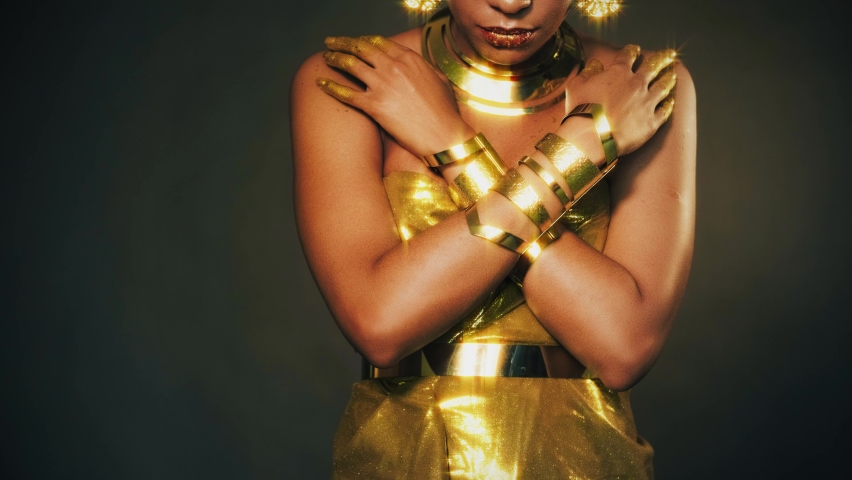 Portrait Closeup fantasy african woman, hand in gold paint. Golden shiny skin dress bracelets necklace earrings arab style. Fashion model sexy girl. metallic makeup lips face buddha statue religion