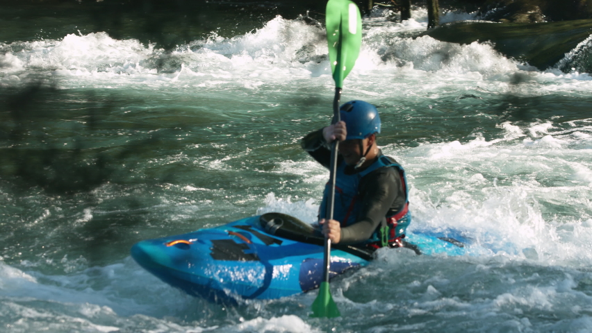 Calm and confident young Caucasian man paddling a blue kayak, skillful maneuvering over river rapids, handheld shot.