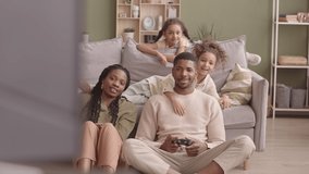 Slowmo of young beautiful Black woman and two playful little girls on sofa watching their father playing video games with controller at weekend