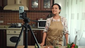 Charming multiethnic, pretty woman, pleasant housewife in a chef's apron, looks at a smartphone camera, mounted on a tripod and records video content for her food blog. Canning. Pickling. Marinating
