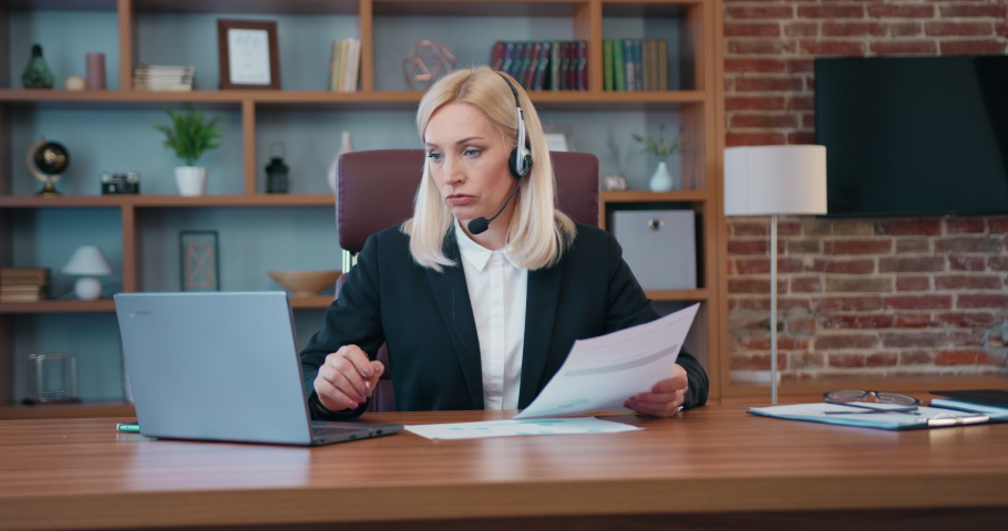 Portarit of charming confident successful blond female office manager with headset which holding business videochat with coworkers on laptop and discussing joint business issues Royalty-Free Stock Footage #1093040037