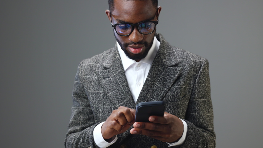 A man African American businessman in glasses holds a phone in his hands looks into the neko and rejoices in a profitable trading deal and receiving income in the office on a gray background | Shutterstock HD Video #1093041695