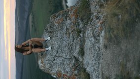 
female tourist sitting on top of a rock in front of an amazing landscape, long hair blowing in the wind.  the girl enjoys the freedom of travel and the beautiful view.  Vertical video