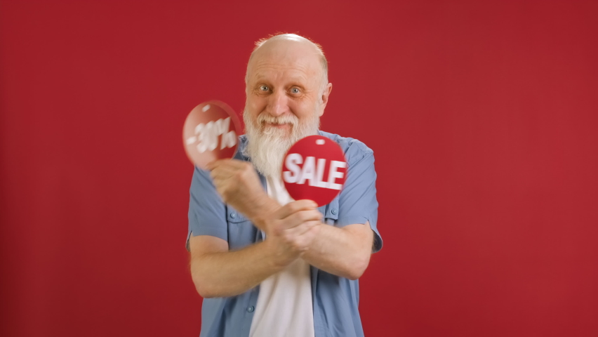 SALE -30 Off. Cheerful old man Dancing and Joyful From Black Friday Sale Holding Two Banners With Inscription SALE and -30 Off Showing Off Inscriptions to Camera on Red Studio Background. | Shutterstock HD Video #1093047395
